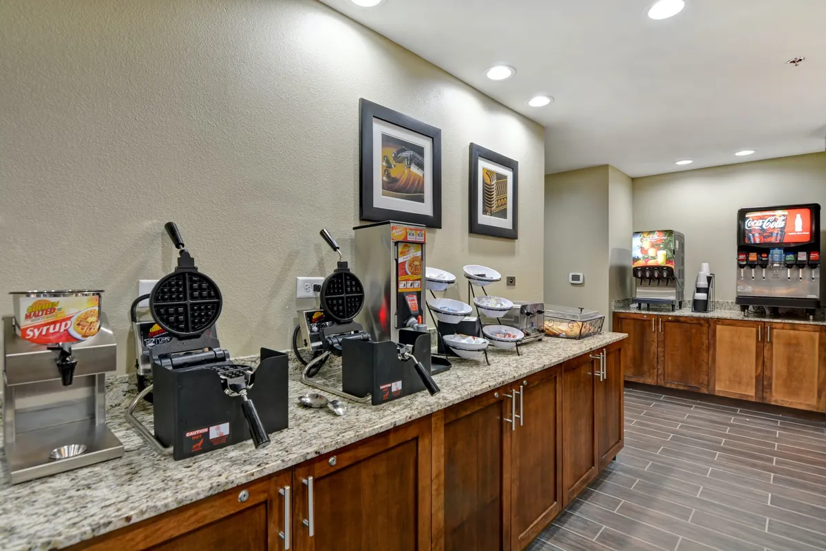 This image showcases a welcoming breakfast setup with waffle makers, a syrup dispenser, a beverage station, and food display bowls, capturing the casual and inviting atmosphere of a complimentary breakfast buffet. The surroundings are bright and encourage socializing, making it easy for families or guests to begin their day together.