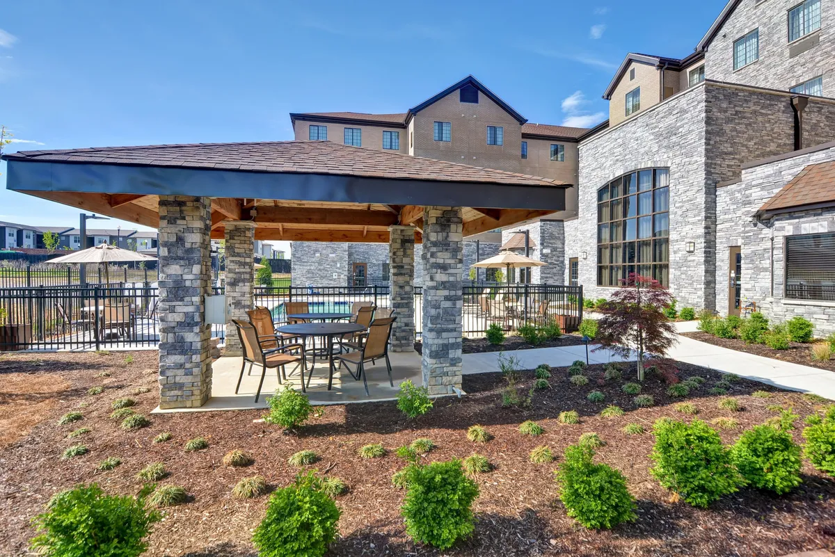 This image captures an inviting exterior scene featuring a stone gazebo with a dining table and chairs, a sparkling pool area, and a well-landscaped garden. The setting is perfect for outdoor dining, relaxing by the pool, or socializing in a tranquil environment.