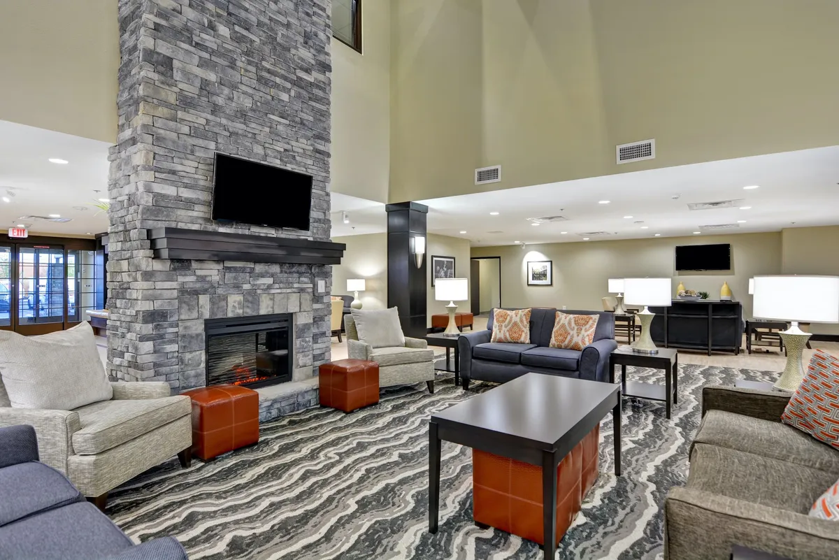 A cozy, inviting living room anchored by a stone fireplace and surrounded by comfortable seating. Large windows offer a view outside, and a textured rug adds warmth to the room. The setup encourages both social gatherings and quiet moments, like watching TV or settling in with a book.