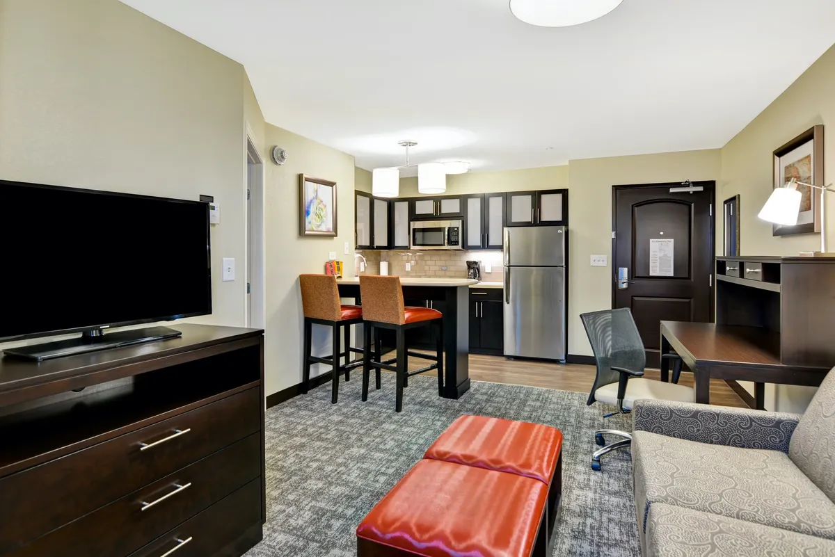 The image showcases a welcoming open-plan living space, featuring a modern kitchen with stainless appliances, a comfortable seating area facing a large television, and stylish artwork on the walls. The scene radiates warmth and functionality, inviting guests to cook, relax, entertain, or catch up on work.