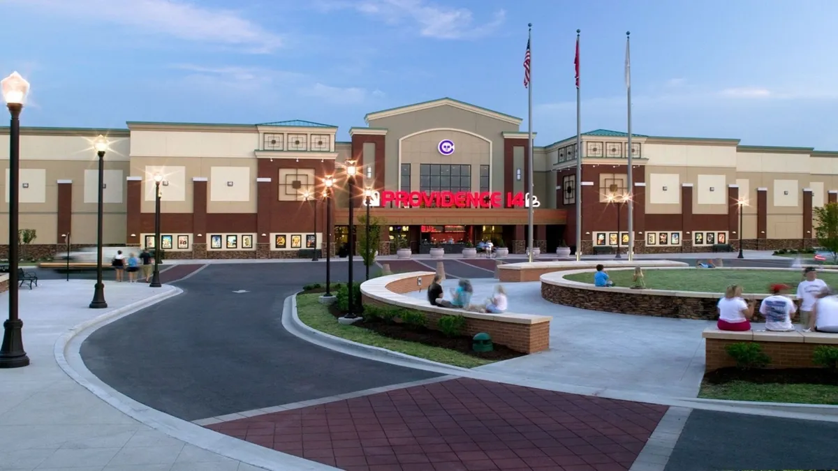 The image showcases the entrance of a modern theater, complemented by a landscaped seating area and illuminated pathway lighting. The lively atmosphere suggests it's an inviting spot for family outings or a relaxed evening after a day of adventure.