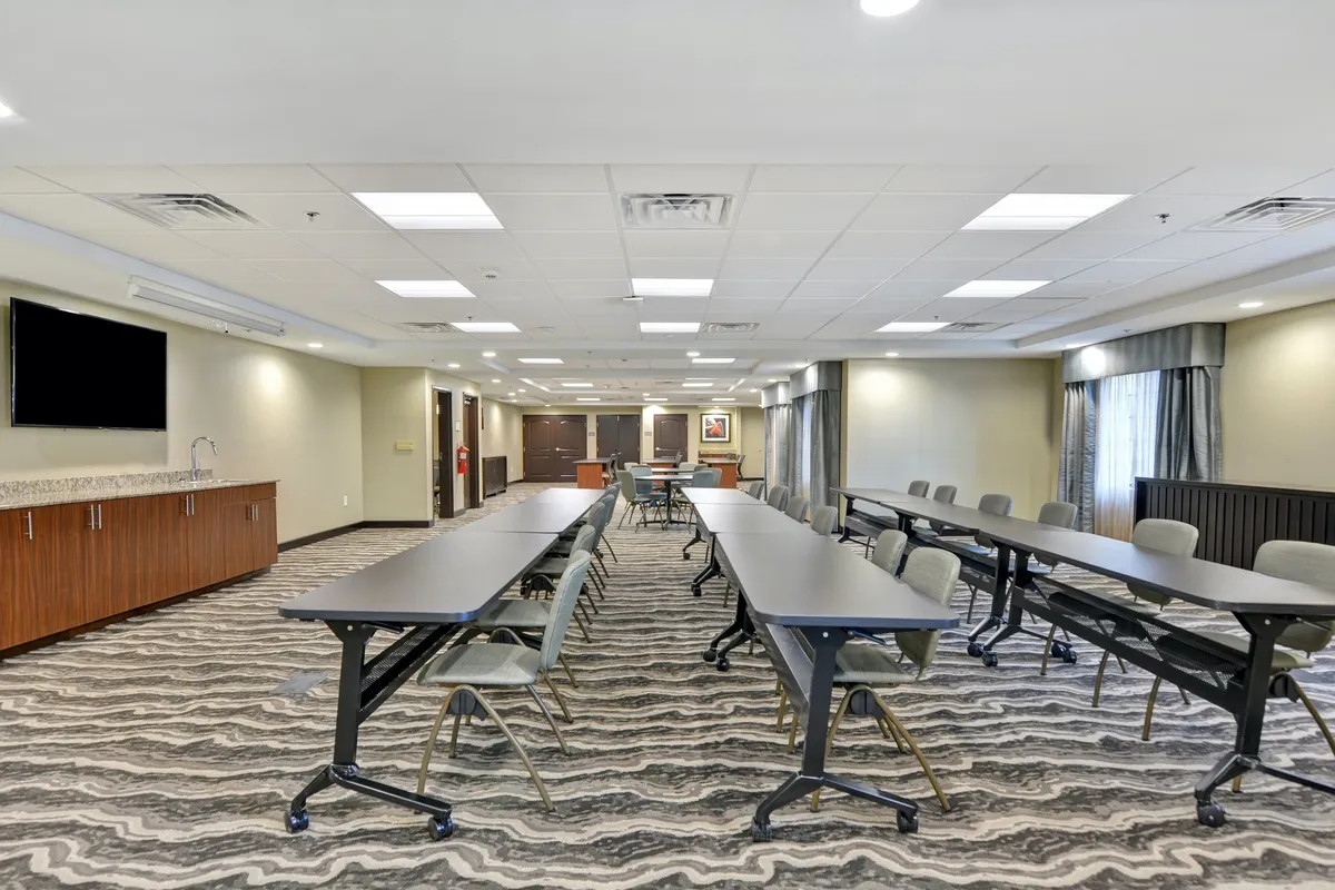 An interior meeting space with long tables arranged in a U-shape, a large wall-mounted screen for presentations, and ample natural light streaming through the windows. The setup is tailored for business meetings, collaborative workshops, and seminars.